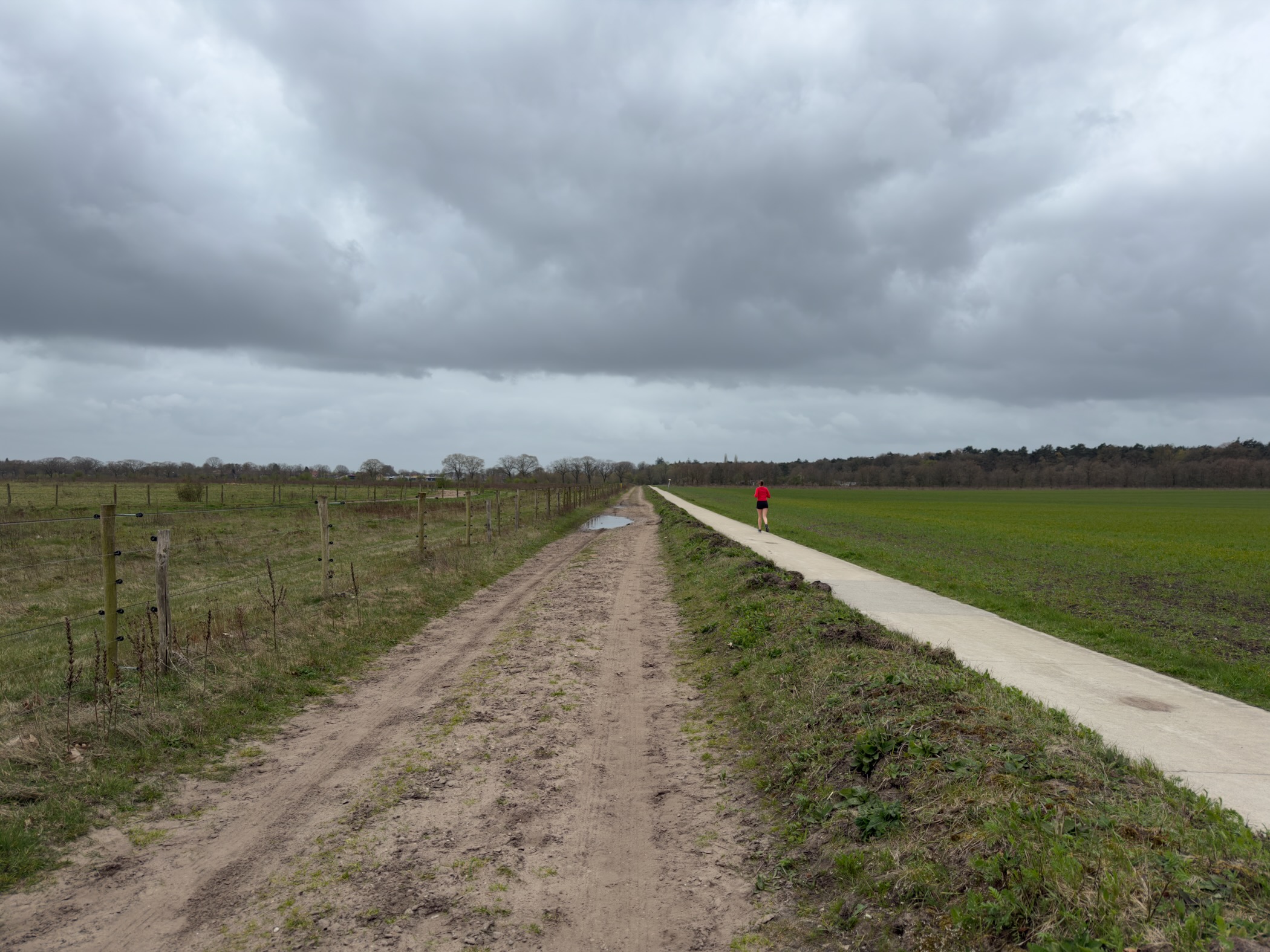 Dirt track and paved path stretching into the distance with a runner in red under grey clouds