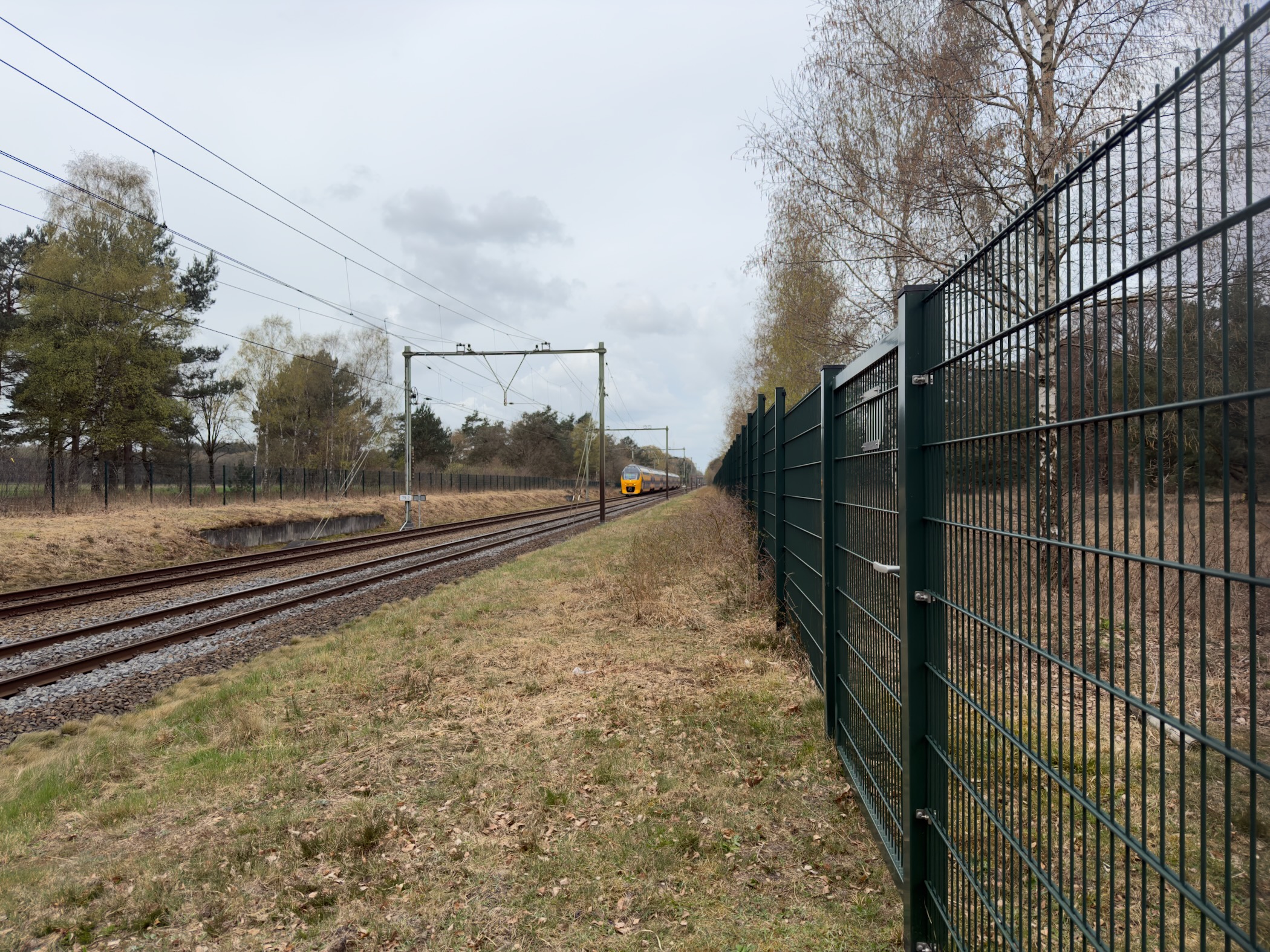 Train approaching along railway tracks beside a green metal fence