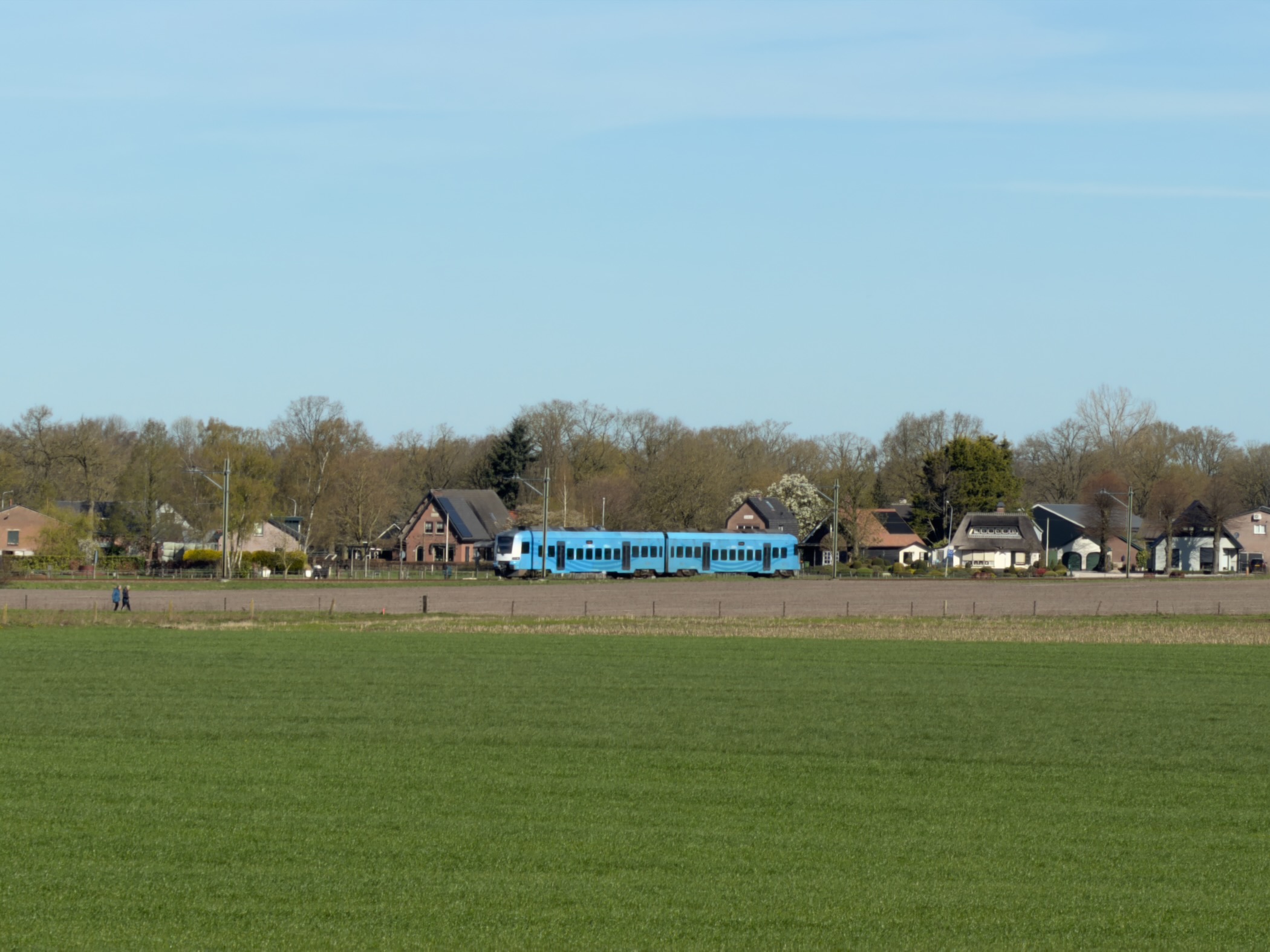 Blue local train passing farmhouses across an open green field