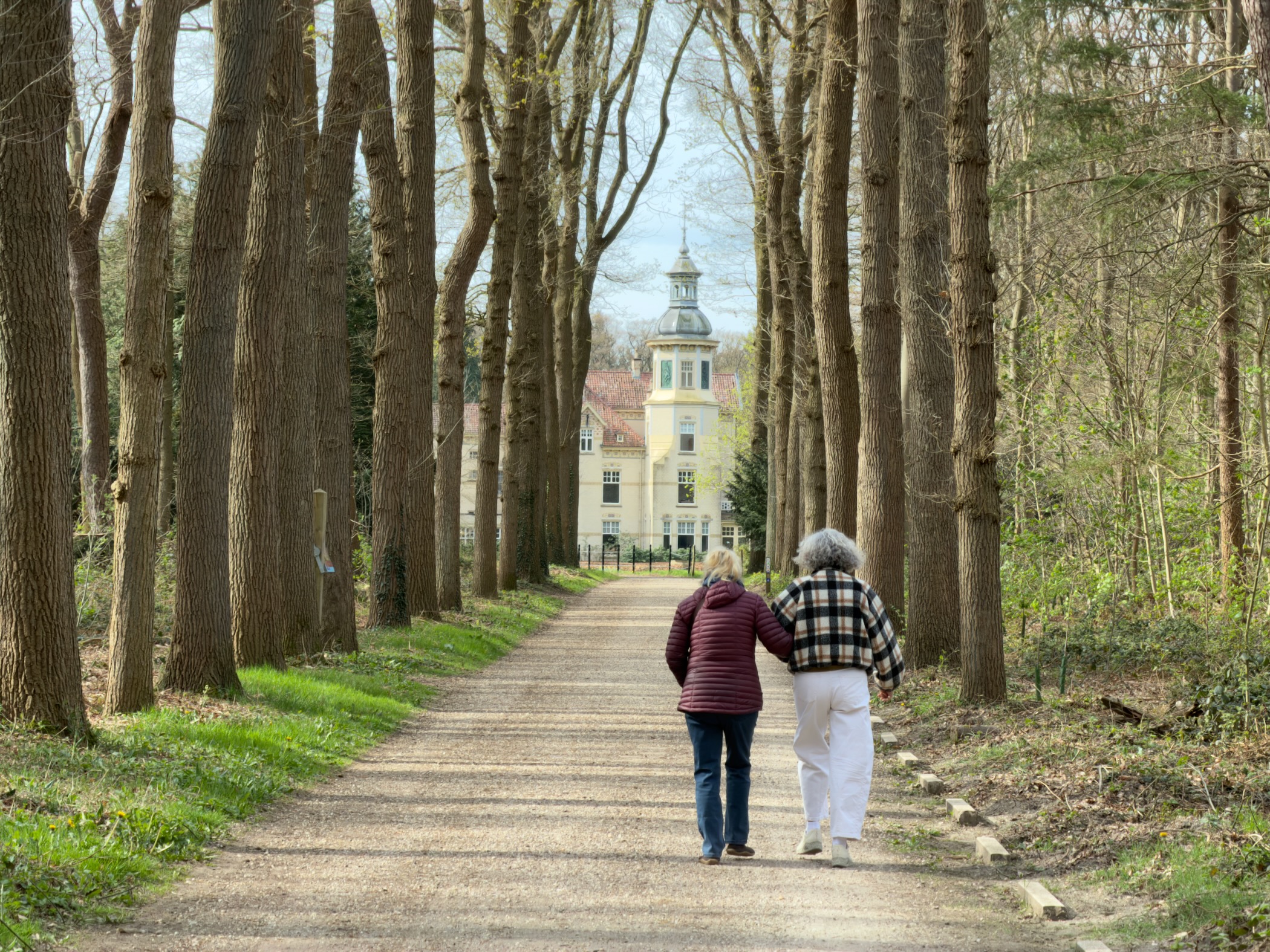 Two walkers on a tree-lined avenue approaching a manor house with a tower