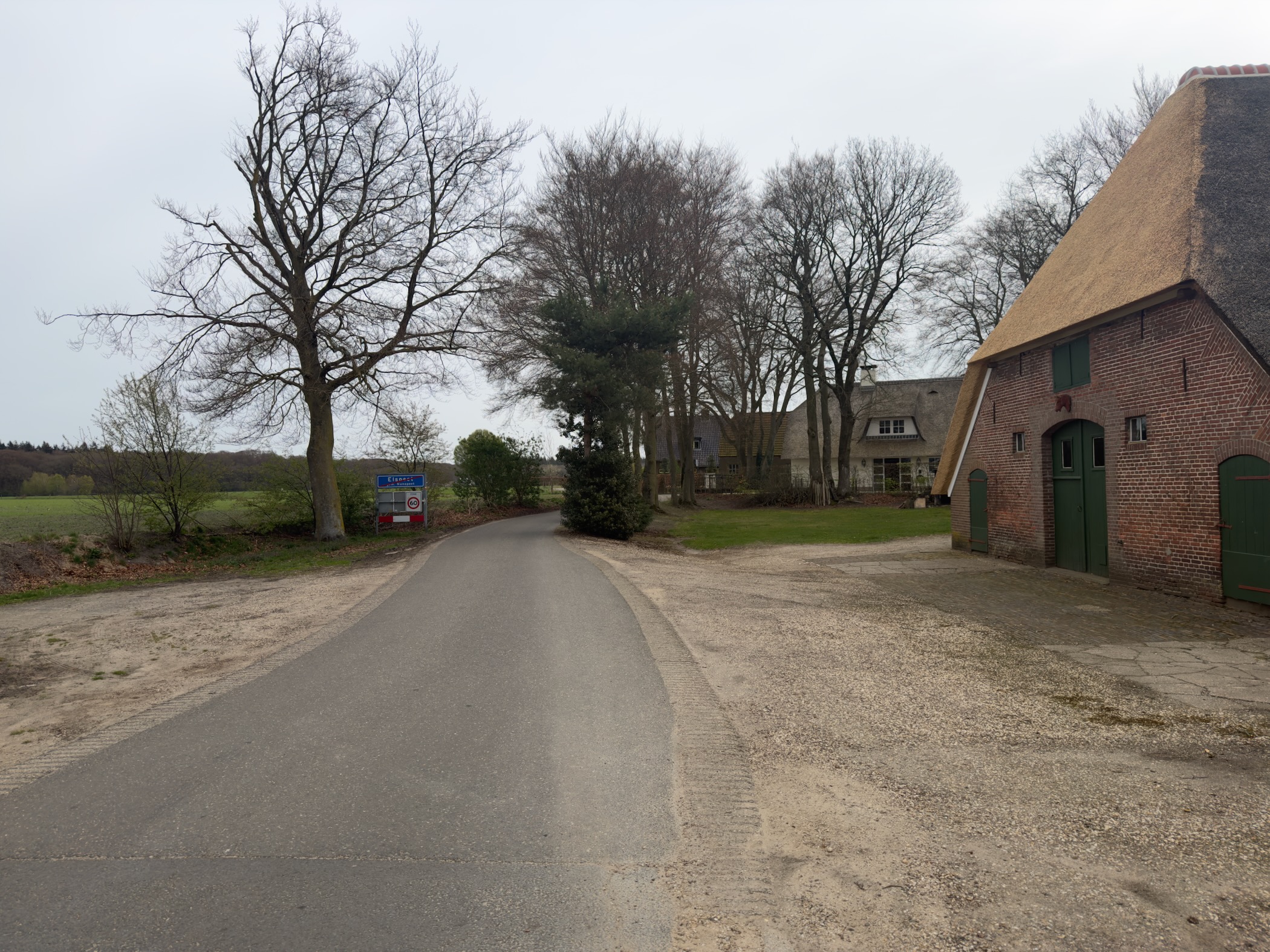 Rural road curving past thatched-roof farmhouses with bare trees under an overcast sky