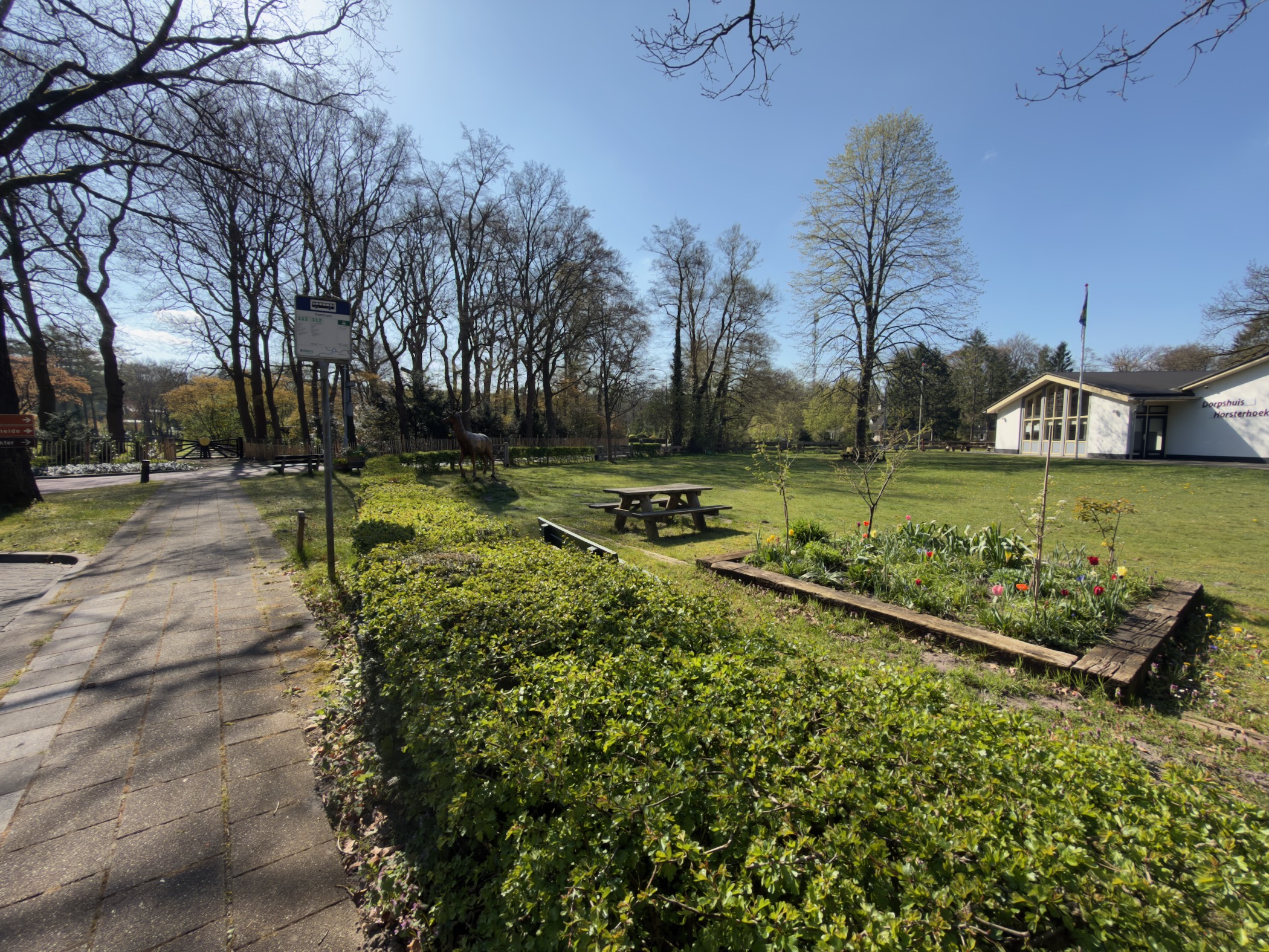 Village green with a picnic bench, flower beds, and a building under blue sky