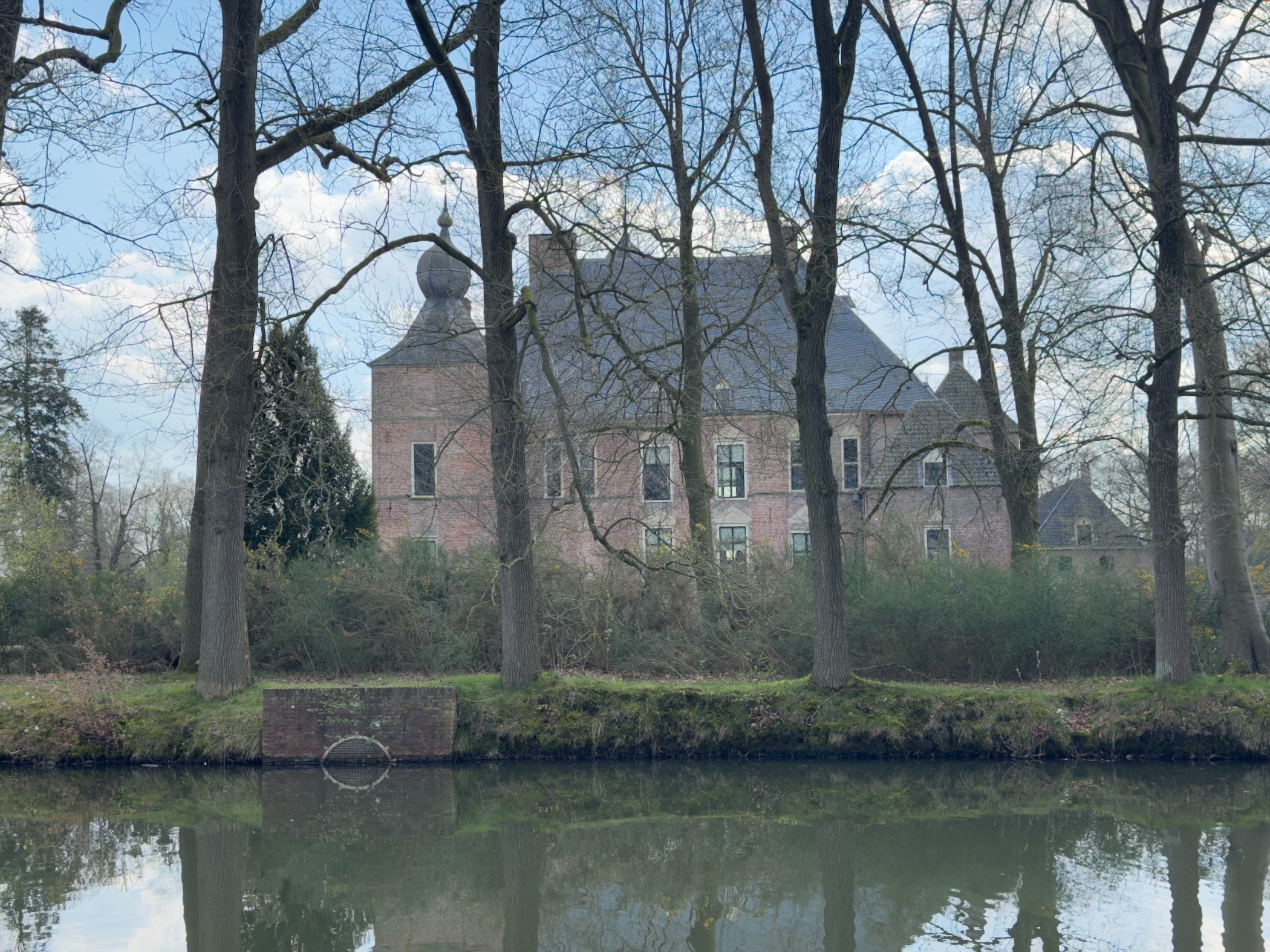 Brick castle with a dome-topped tower seen across a calm moat through bare trees