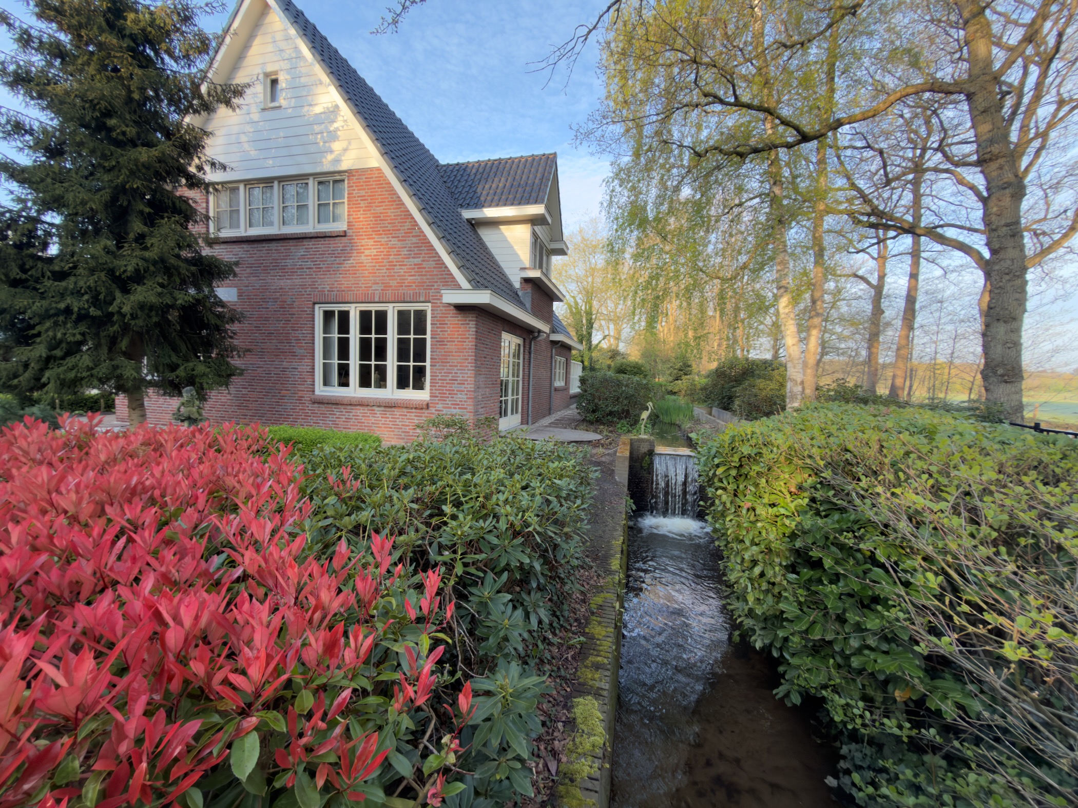Brick house with a stream flowing alongside it and bright red photinia bushes in the foreground