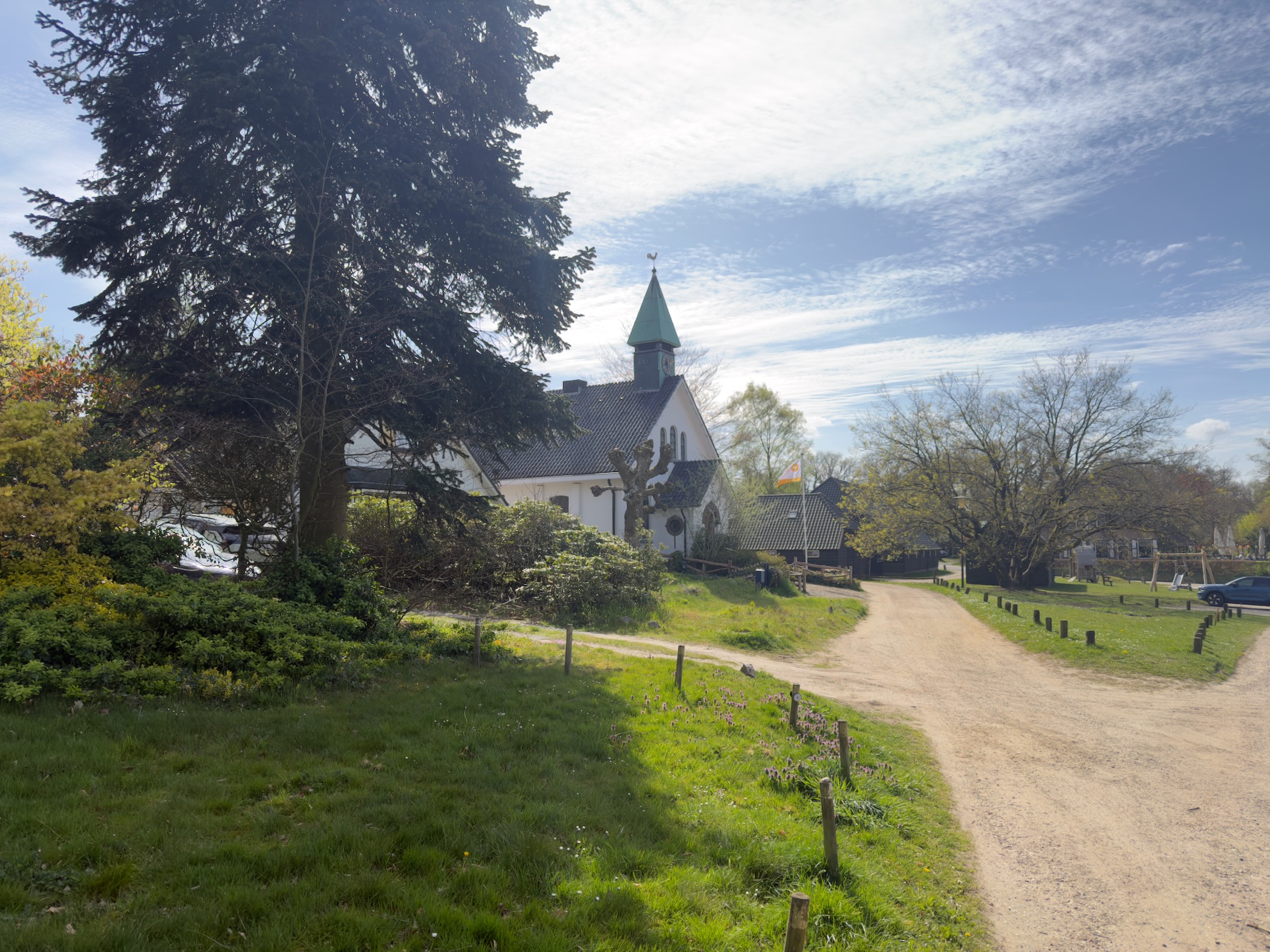 White chapel with green spire in Hoog Soeren village, seen from a dirt track lined with grass and trees