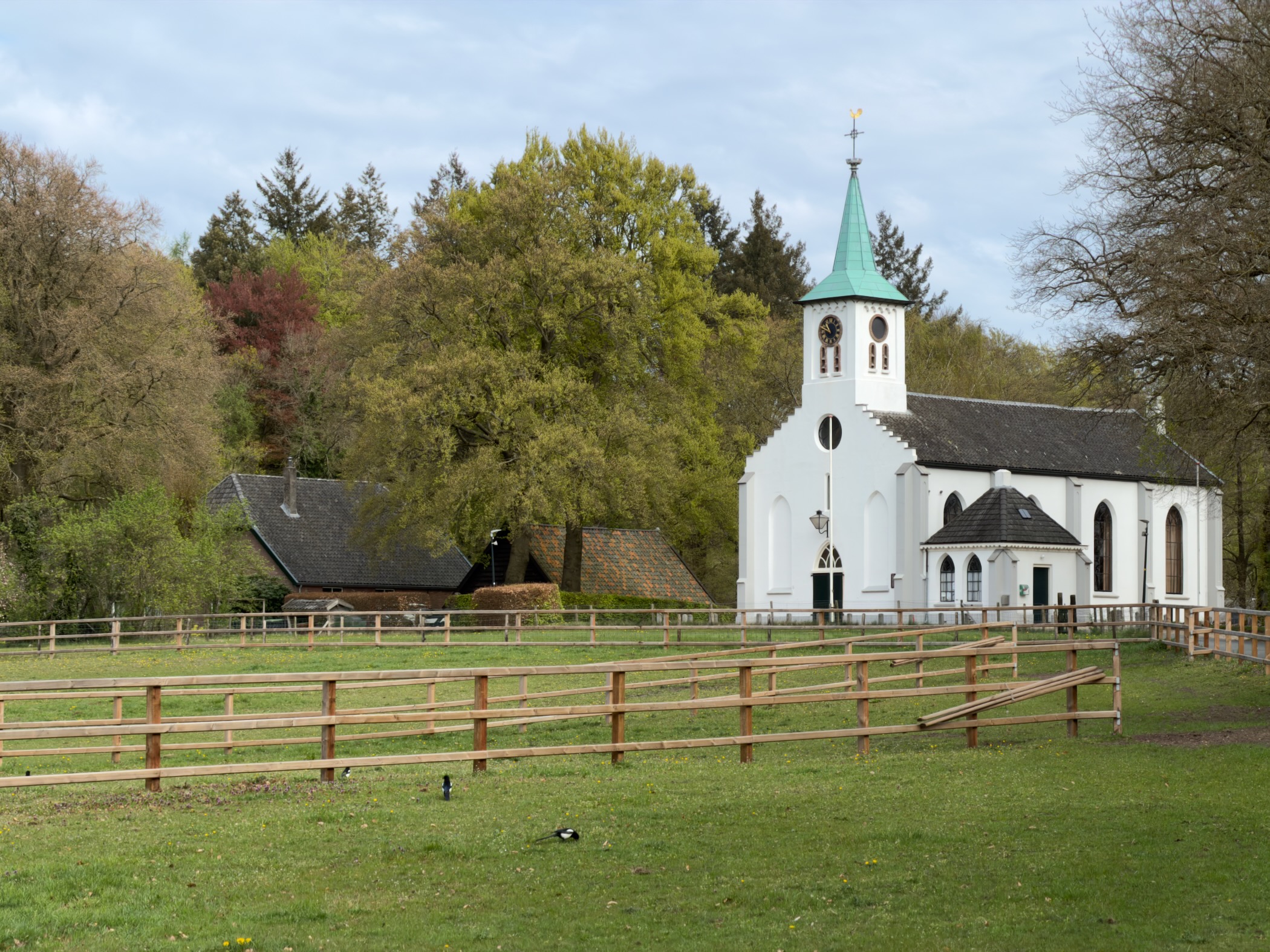 White church with green spire next to a small thatched barn behind a wooden fence