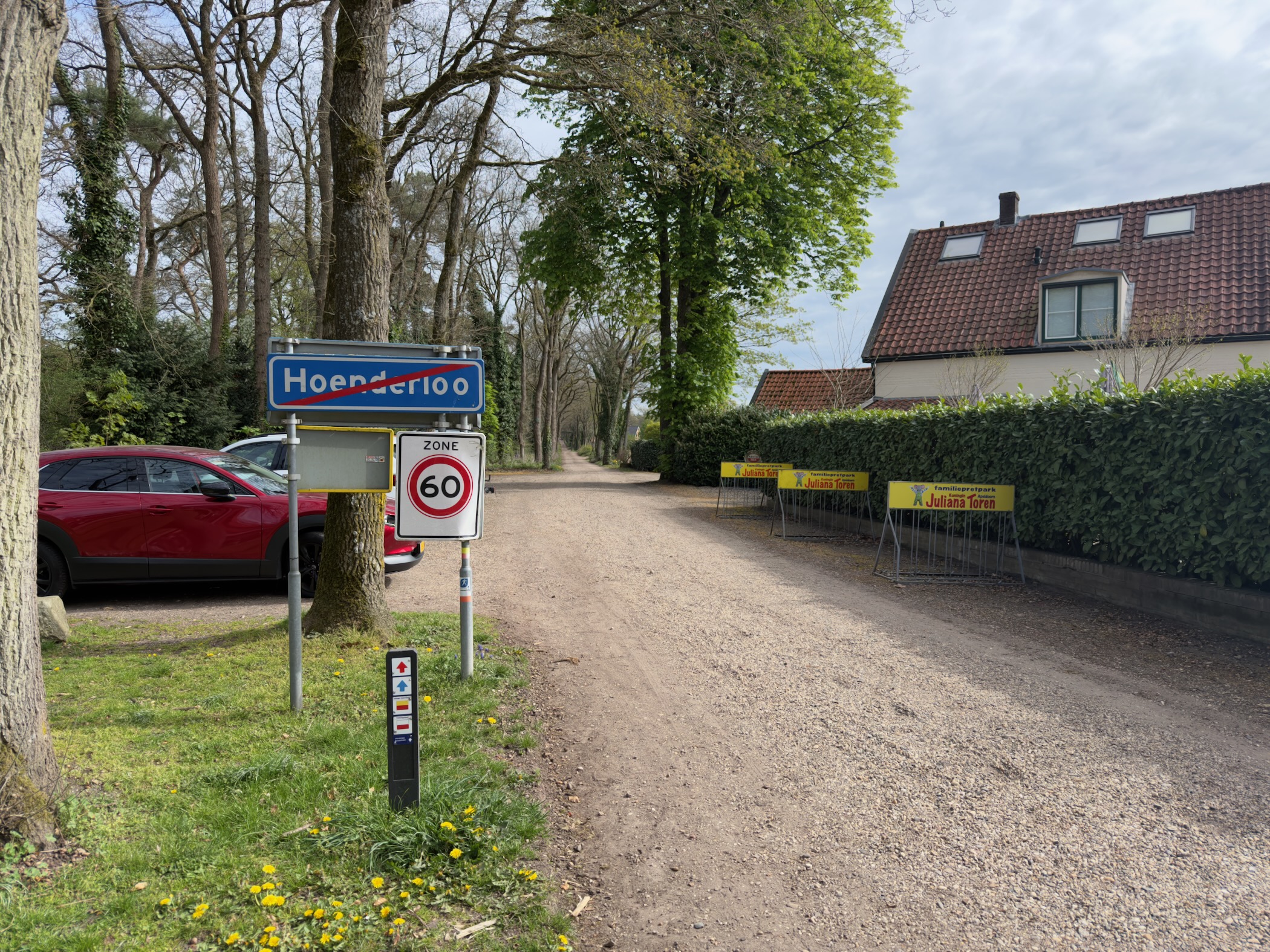 Hoenderloo town sign beside a sandy road with a brick house and road-work barriers