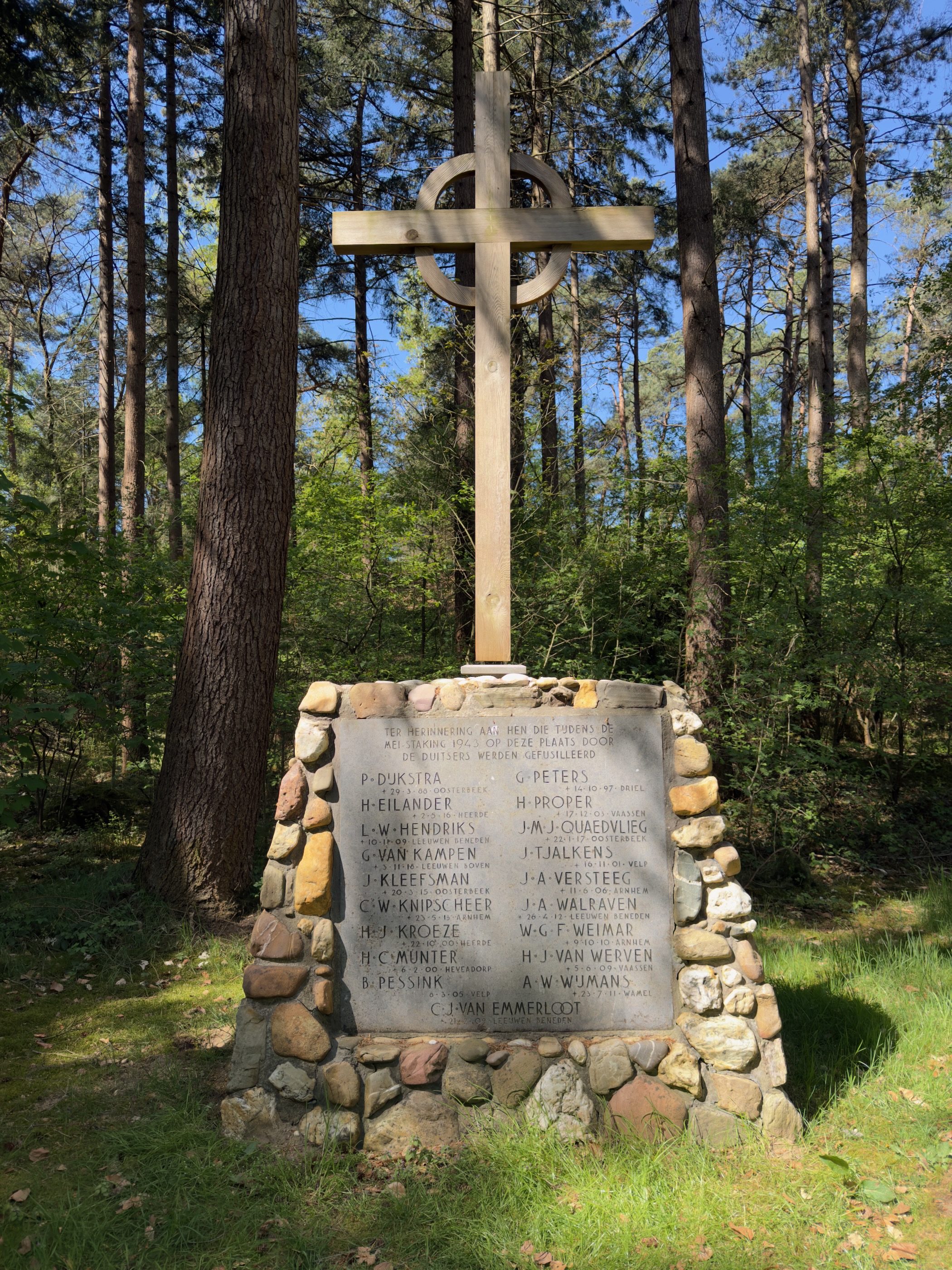 Stone war memorial topped with a wooden cross, engraved names on a plaque in the forest