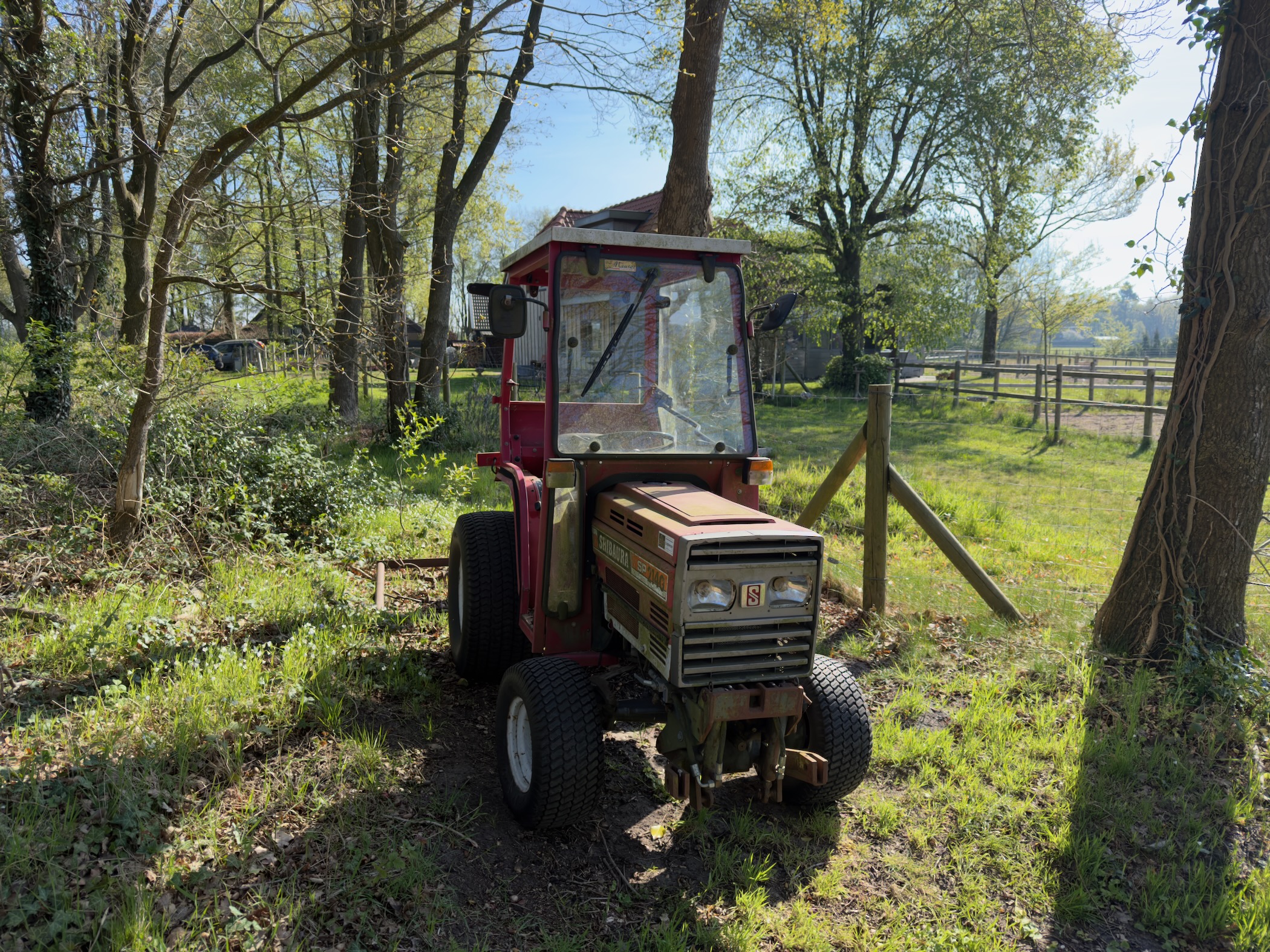Old red tractor parked beside a wooden fence in a farmyard clearing