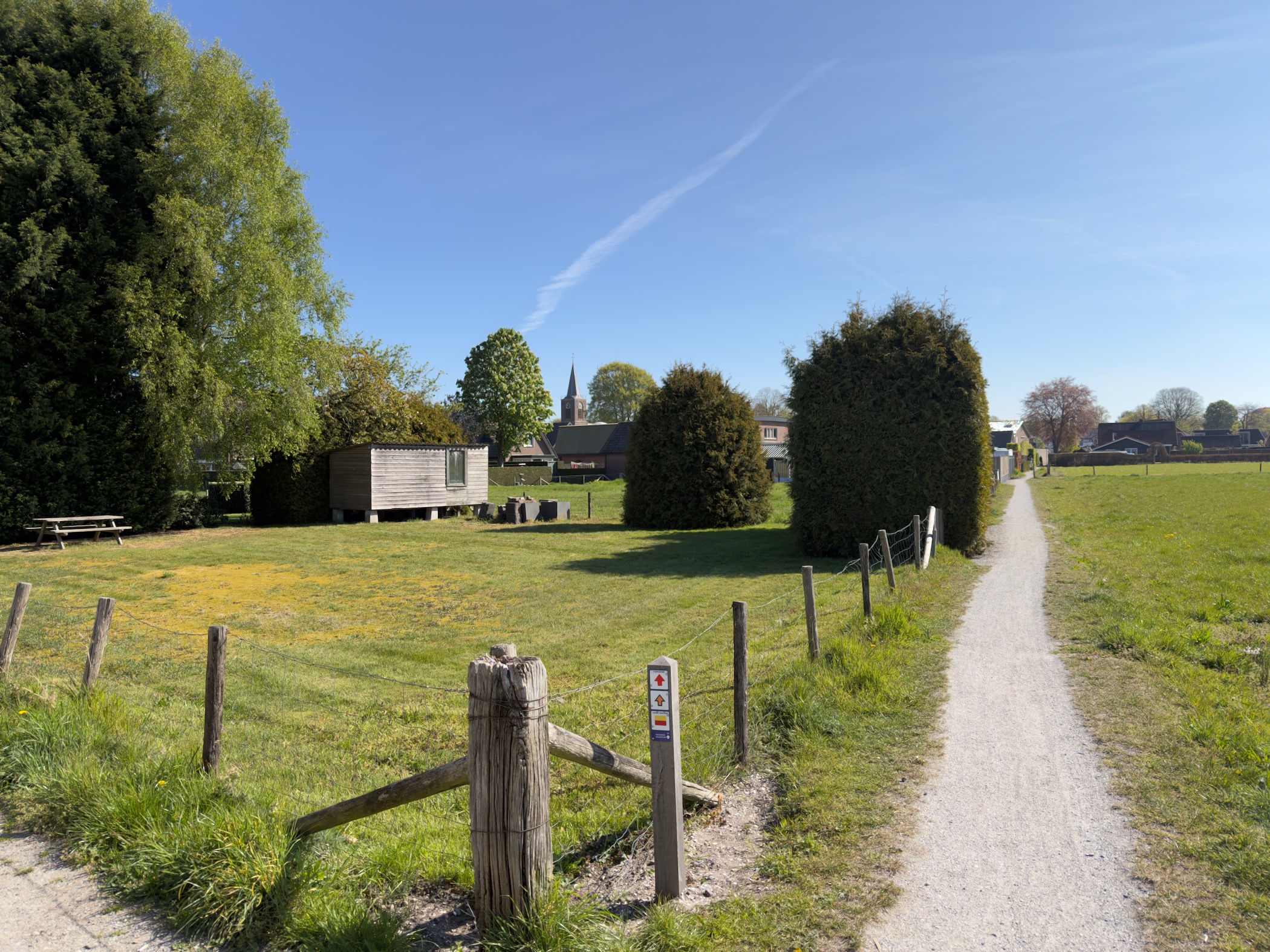 Grass path across a lawn with a village church spire rising behind the trees