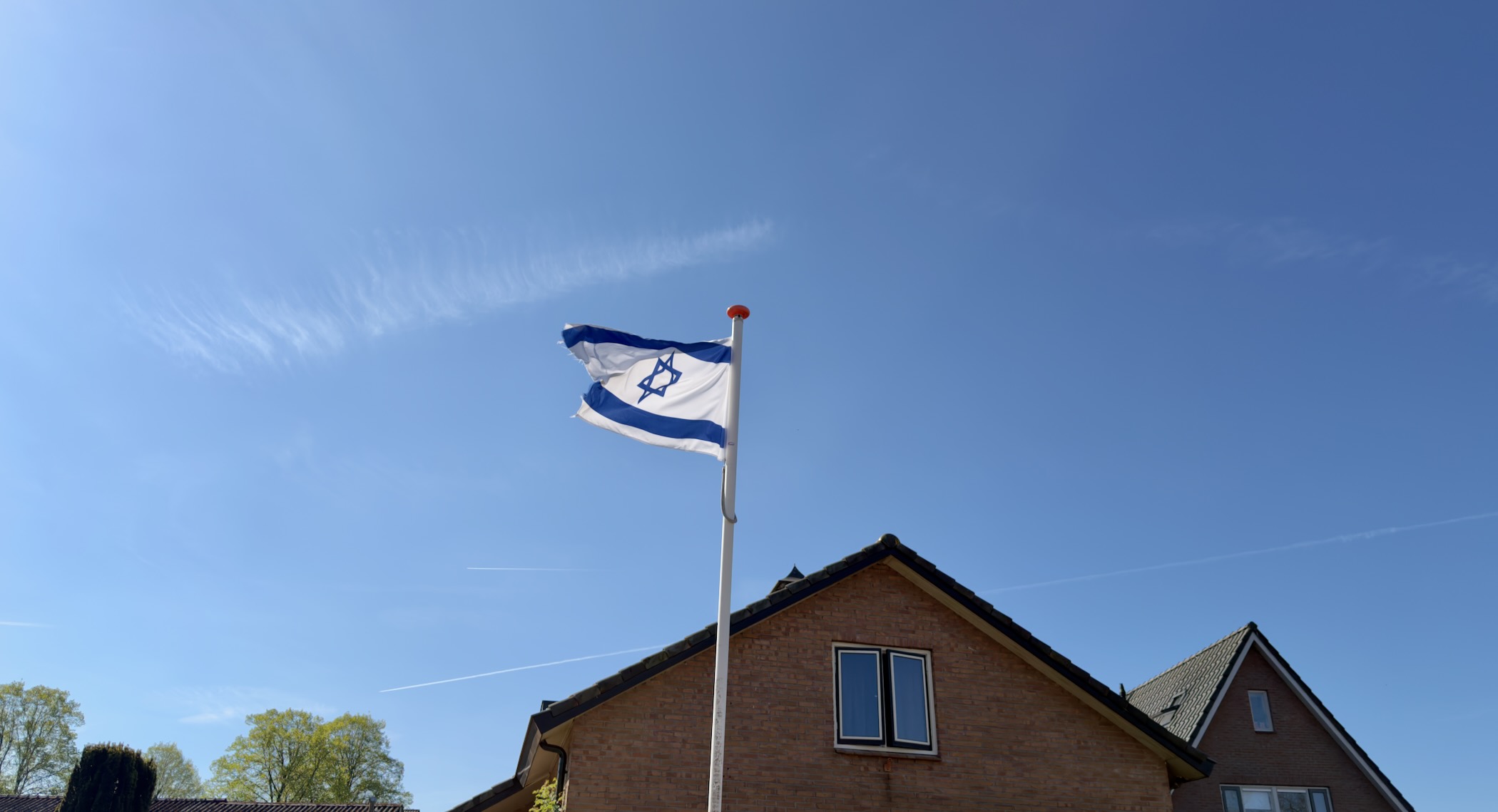 Israeli flag flying from a pole above a brick gable-roofed house under a clear blue sky